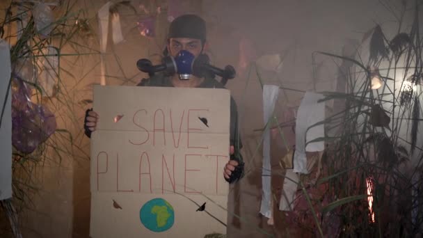 言葉でポスターを保持しているガスマスクの男は惑星を救う プラスチックごみを背景に燃焼させます 環境問題の概念 プラスチックごみ 不法投棄 廃棄物焼却 — ストック動画