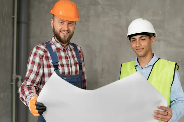 幸せな建築家や建設エンジニアや測量士の議論の計画や設計図 2人の男が硬い帽子をかぶっていて 屋内の家の建設現場に立っている カメラを見ると — ストック写真