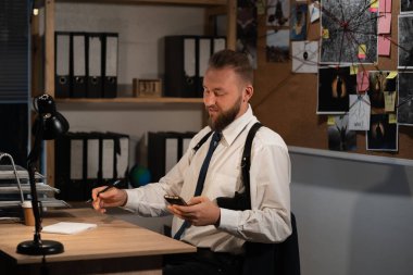 Profesyonel bir dedektif gece ofiste bir cinayet üzerinde çalışıyor. Masada oturuyor, elinde bir akıllı telefon tutuyor ve internetten haberleri okuyor. Boşluğu kopyala.