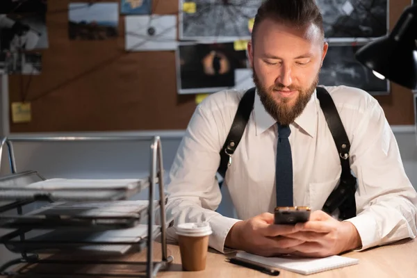 A male detective is sitting in the office flipping through the news feed. A detective board with photos, a map and clues connected by a thread on the wall. copy space.