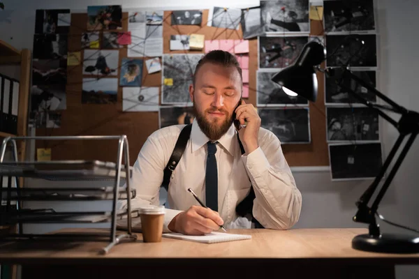 A professional detective works on a murder in the office at night. Sitting at the table takes a call from a witness and writes down the testimony in a notebook. copy space.
