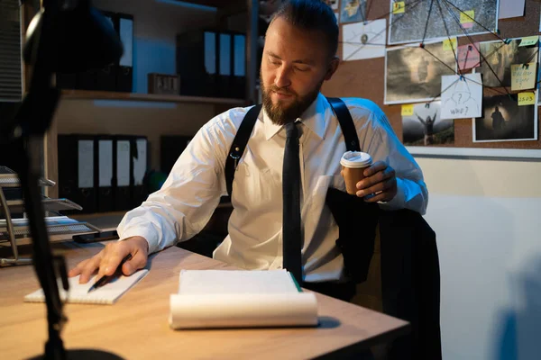 A professional detective is working on a murder in the office at night. He sits at the table with coffee in his hands and reads notes in a notebook. copy space.