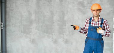 Gri duvarda oturan, müteahhit üniforması giyen, kasklı, gülümseyen, gülümseyen bir gülümsemesi olan genç bir Arap. Pankart