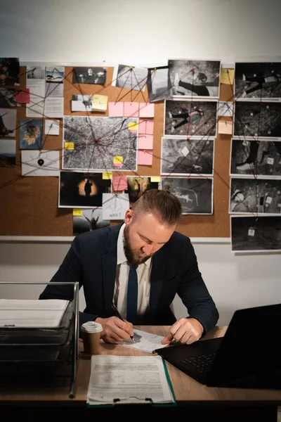 Detective working at desk in his office, evidence board on background, copy space