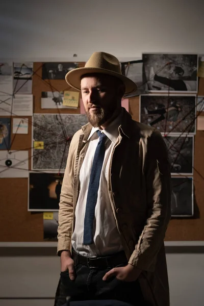 Portrait of pensive private detective in trench coat and hat near evidence board in office at night in the office.