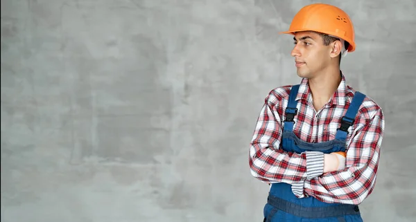 Portrait of confident arabic builder in overall and shirt having arms crossed, looking at side, isolated on gray wall background, indoors. house renovation, repair concept. Banner