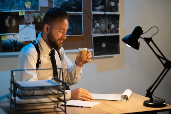 Detective working in the office at night drinking coffee, evidence board on the background of the police
