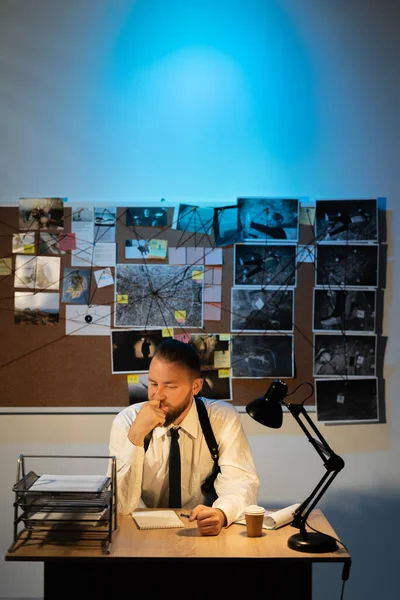 Detective thinking working at desk in his office at night, evidence board on background