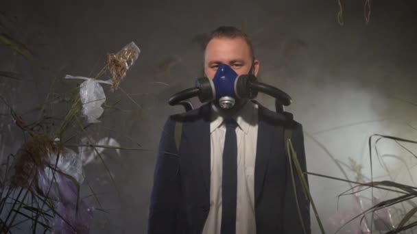 身穿夹克的男子站在烟雾弥漫的环境中 手里拿着一个停止化学战的标志 氧气罐与呼吸面罩相连 干草上的塑料废物 生态灾害概念 — 图库视频影像