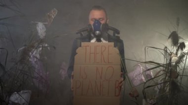 Oksijen maskeli beyaz sakallı bir adam gaz dolu bir balonla plastik atıkla kaplı kuru otların arasında B gezegenine ait bir işaret tutmuyor. Çevre felaketi kavramı