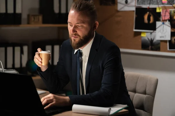 Detective working in the office using a computer, process of analyzing the evidence of a criminal case, investigator drinks coffee at night in the workplace