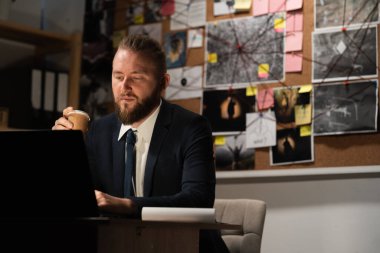 Dedektif gece ofiste bilgisayar kullanarak çalışıyor, işyerinde kahve içiyor, arka planda kanıt panosu var.