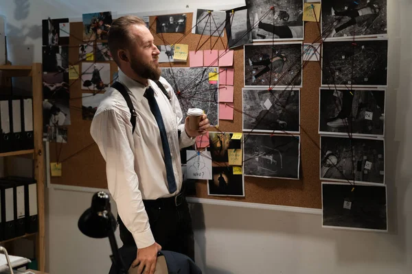 Detective looking at evidence board in office, investigator drinking coffee at night on workplace, copy space