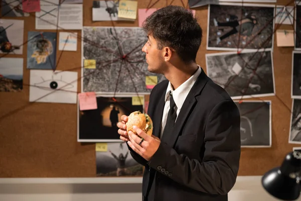 Detective looking at evidence board in office and eating fast food hamburger, workplace food concept
