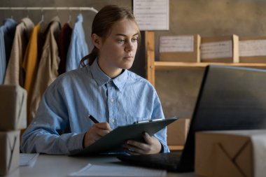 İnternetten yeni kıyafetler satıyorum. Genç bayan küçük işletme sahibi, karton kutularda nakliye edilen moda kıyafetleri için dizüstü bilgisayarda çalışıyor. Boşluğu kopyala
