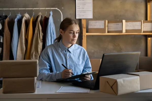 Online store selling new clothes from home. Young woman small business owner working on laptop shopping for fashion clothing shipped in shipping cardboard boxes. Copy space