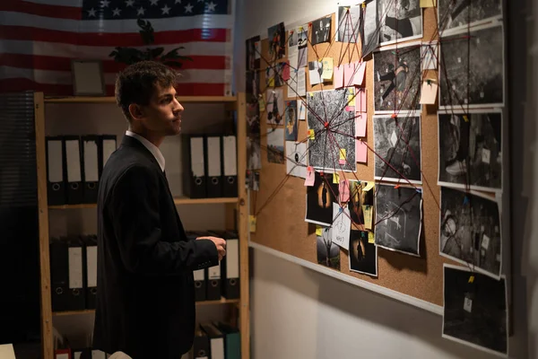 Detective looking on investigation board with photos of suspected criminals, crime scenes and evidence with red threads, working in the office. Copy space