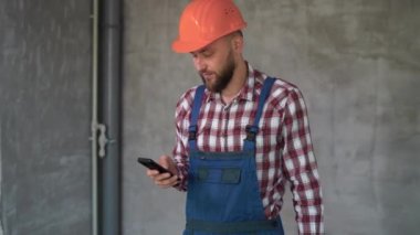 Sakallı işçi ya da kasklı müteahhit gri duvar arkaplanı üzerinde akıllı telefon kullanıyor. Meslek, inşaat ve onarım kavramı
