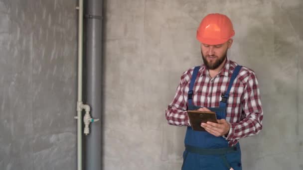 在灰墙背景下持有数字平板电脑的建筑工人或修理工 头戴橙色头盔的工头正在检查项目的进度 建筑及建筑概念 — 图库视频影像