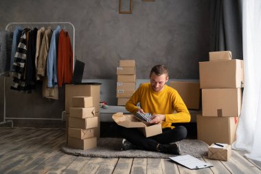 Genç adam evdeki kutuya kumaş paketleyen küçük bir işletme sahibi kuruyor, serbest satıcı çevrimiçi dükkanda paketleme işlemi için ürün hazırlıyor. Küçük işletme kavramı