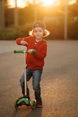 Uzun sarı saçlı, kot pantolon giyen ve kırmızı kapüşonlu güzel bir çocuk güneşli bir yaz gününde scooter sürüyor..