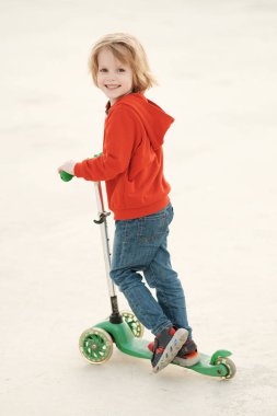 Uzun sarı saçlı, kot pantolon giyen ve kırmızı kapüşonlu güzel bir çocuk güneşli bir yaz gününde scooter sürüyor..