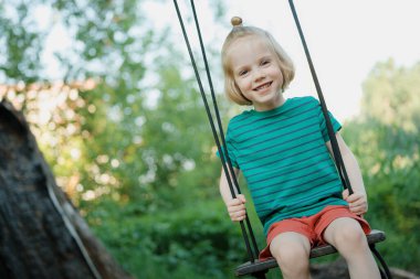 Güneşli bir yaz gününde, uzun sarı saçlı tatlı çocuk temiz havada sallanıyor. Yeşil tişört ve kırmızı şort giyen çocuk.