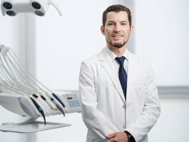 Healthcare, profession, stomatology and medicine concept - smiling and confident male middle aged dentist wearing white coat stand with hands crossed near medical equipment in a dental studio