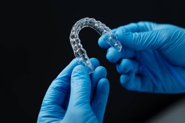 Invisible orthodontics cosmetic aligners in hands of doctor wearing sterile gloves isolated on a black background, tooth aligners, plastic braces. Modern teeth retainers created on a 3d printer.