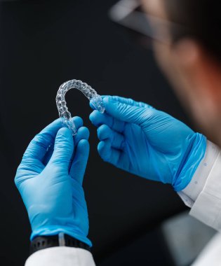Invisible orthodontics cosmetic aligners in hands of doctor wearing sterile gloves isolated on a black background, tooth aligners, plastic braces. Modern teeth retainers created on a 3d printer.