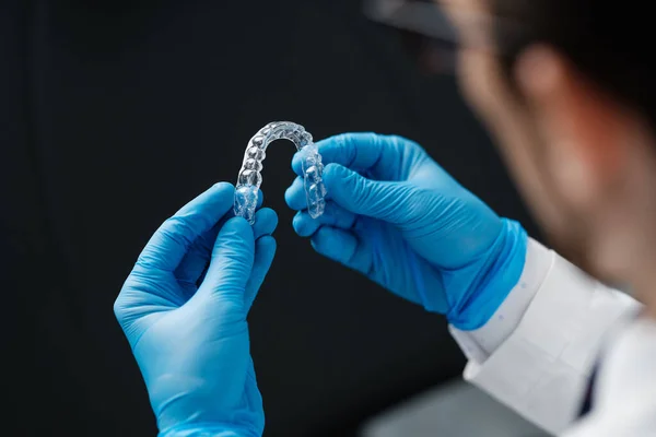 Invisible orthodontics cosmetic aligners in hands of doctor wearing sterile gloves isolated on a black background, tooth aligners, plastic braces. Modern teeth retainers created on a 3d printer.