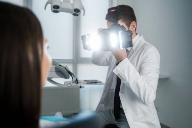 Handsome dentist making photo of female patients smile after treatment in dental office. Special camera with flash ring shadowless.