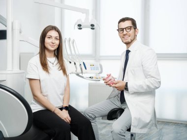 Happy male dentist wearing white coat sitting near dental unit with caucasian female patient with a smile looking at camera in dental office