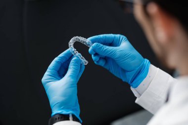 Invisible orthodontics cosmetic aligners in hands of doctor wearing sterile gloves isolated on a black background, tooth aligners, plastic braces. Modern teeth retainers created on a 3d printer.