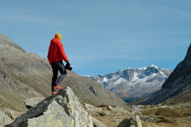 Back view of a tourist man in red jacket and camera standing with a view on Julier Pass, Switzerland clipart