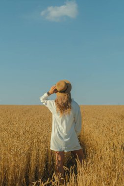 Back view of young woman in a white dress and a straw hat on a wheat field in the summer. The concept of outdoor recreation, a trip to the village