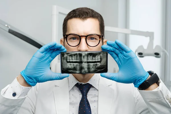 Male dentist cover his mouth with a smartphone with X-ray of his teeth on a screen of device. Modern orthodontist in dental clinic concept