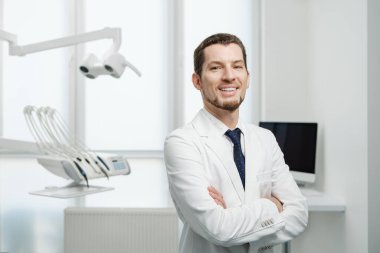 Healthcare, profession, stomatology and medicine concept - smiling and confident male middle aged dentist wearing white coat stand with hands crossed near medical equipment in a dental studio