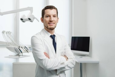 Healthcare, profession, stomatology and medicine concept - smiling and confident male middle aged dentist wearing white coat stand with hands crossed near medical equipment in a dental studio