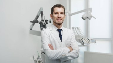 Healthcare, profession, stomatology and medicine concept - smiling and confident male middle aged dentist wearing white coat stand with hands crossed near medical equipment in a dental studio