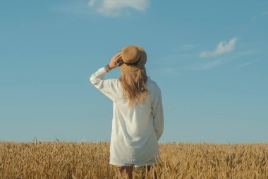 Back view of young woman in a white dress and a straw hat on a wheat field in the summer. The concept of outdoor recreation, a trip to the village