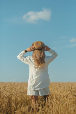 Back view of young woman in a white dress and a straw hat on a wheat field in the summer. The concept of outdoor recreation, a trip to the village