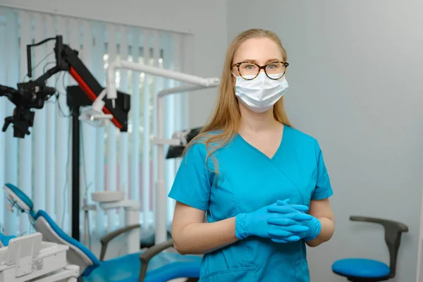 Happy caucasian female dentist in blue lab coat and protective facial mask posing with pretty smile looking at camera in her dental office