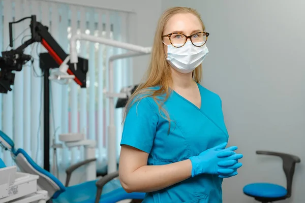 Happy caucasian female dentist in blue lab coat and protective facial mask posing with pretty smile looking at camera in her dental office
