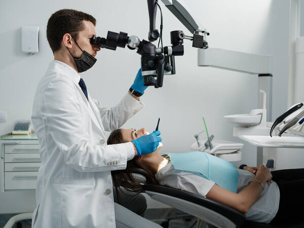 Male dentist using dental microscope treating female patient teeth at dental clinic office. Medicine, dentistry and health care concept. Dental equipment