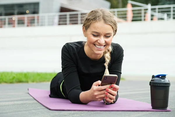 金髪のきれいな女性が手にスマートフォンとフィットネスラグに横たわっています 屋外での訓練後のチャット — ストック写真