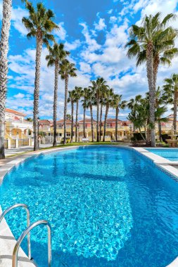 Boş yüzme havuzu etrafında dikey palmiye ağacı. İçinde mavi su olan yerleşim yerlerinin içinde modern lüks evler var. Güneşli yaz gününde bulutlu gökyüzüne karşı. Costa Blanca, Alicante, İspanya