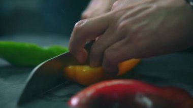 Unknown male chef hands cutting yellow, green and red fresh raw paprika vegetables for slices, close up shot of arms and worktop. Restaurant workflow in kitchen