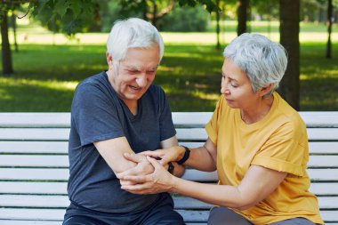 During morning sportive stroll or making exercises in a park, elderly 60s man got injured his shoulder gripping arm sit in bench with caring disappointed wife. Traumas, injures of older people concept