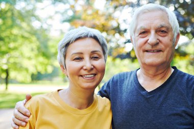 Optimistic active old 60s couple in love pose outdoor, smile looking at camera during a morning walk in a public park. Happy endless marriage, romantic relations, older family portrait concept